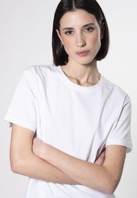 Camiseta blanca de algodón con mangas cortas y escote redondo. Textura simple y suave, sin patrones ni acentos visibles. El modelo tiene los brazos cruzados.
