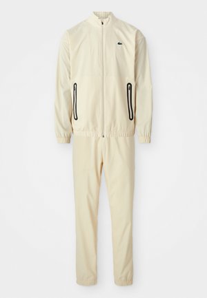 Ensemble de survêtement couleur crème avec une veste zippée et un pantalon assorti. Comprend des poches zippées noires et un logo vert sur la veste.
