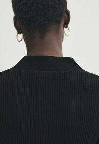 Schwarzer gerippter Pullover mit hohem Kragen, der ein strukturiertes Strickmuster aufweist. Goldene Creolen betonen das schlichte Design.