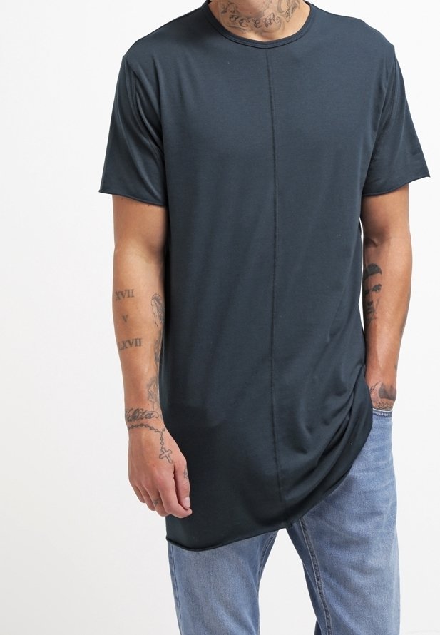 Homme portant un t-shirt bleu foncé à ourlet asymétrique et un jean bleu clair avec des tatouages visibles sur les bras et le cou, debout les mains détendues.