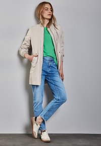 Helle beige Bomberjacke, grünes T-Shirt, hellblaue Jeans und weiße Sneakers mit grauen Akzenten; Model posiert vor einem neutralen Hintergrund.
