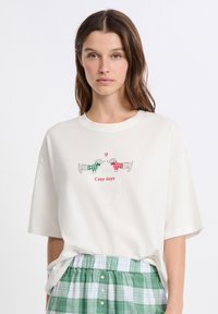 Kobieta nosząca białą koszulkę z dwoma jamnikami w świątecznych swetrach, które stoją naprzeciwko siebie, oraz tekstem "Przytulne dni" na czerwono.