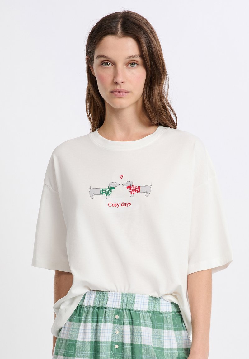 Kobieta nosząca białą koszulkę z dwoma jamnikami w świątecznych swetrach, które stoją naprzeciwko siebie, oraz tekstem "Przytulne dni" na czerwono.