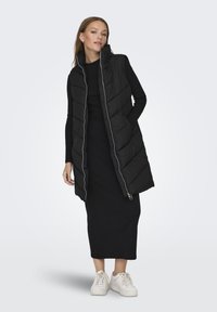 Gilet en duvet noir avec un design matelassé vertical, col montant et accents de fermeture éclair argentés. Porté par-dessus une robe noire avec des baskets blanches.