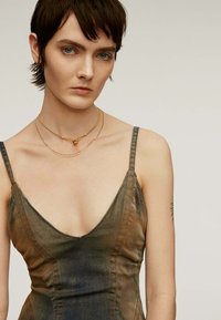 Vestido de denim con un profundo escote en V, que presenta tonos degradados de marrón y azul, y costuras naranjas en contraste. Dos collares de oro superpuestos.