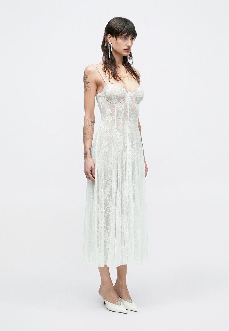 Robe en dentelle transparente avec broderies florales, bretelles spaghetti et silhouette fluide ; portée avec des mules blanches pointues, mettant en valeur la texture légère.