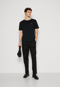 T-shirt noir à col rond avec un logo simple, associé à un pantalon noir ajusté. Le modèle tient un sac noir et porte des chaussures noires.