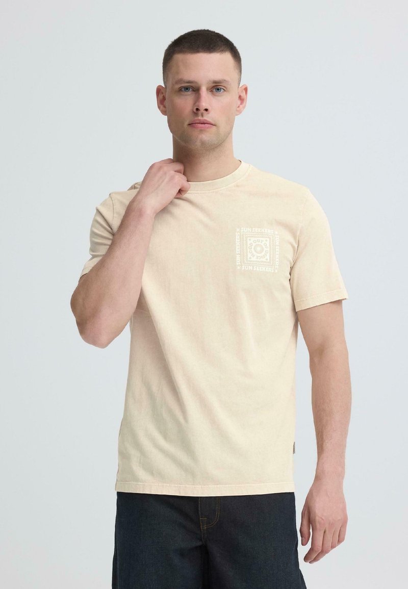 Άνδρας με κοντά μαλλιά που φοράει ανοιχτό μπεζ μπλουζάκι με λευκό τετράγωνο σχέδιο στο στήθος και σκούρα μπλε τζιν, στέκεται μπροστά σε απλό φόντο.