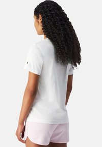 Hvid bomulds t-shirt med korte ærmer, med et lille broderet logo på ærmet. Båret med lyserøde shorts.