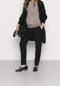 Graviditetsoutfit med en mönstrad topp i ljusrosa och svart, en lång svart kofta, svarta byxor och svarta loafers med guldaccenter.