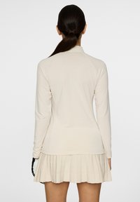 Hellbeige Langarm-Polohemd mit weicher Textur, kombiniert mit einem gefalteten Rock in passender Farbe, der schlichte Linien und Formen zeigt.