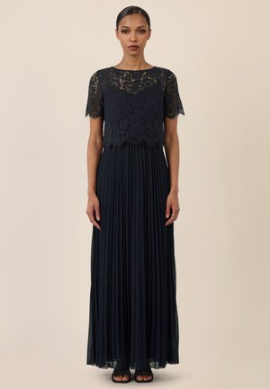 Frau mit zurückgebundenem Haar, die ein schwarzes Spitzenoberteil mit kurzen Ärmeln und einen langen, plissierten Rock trägt, steht vor einem beigen Hintergrund.