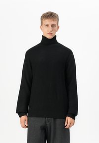 Jongen met kort haar, gekleed in een zwarte gebreide coltrui en donkergrijze broek, staand tegen een witte achtergrond.