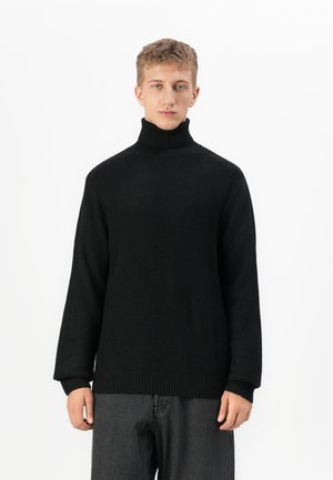 Joven con pelo corto que lleva un suéter negro de cuello vuelto de punto y pantalones gris oscuro, de pie contra un fondo blanco.