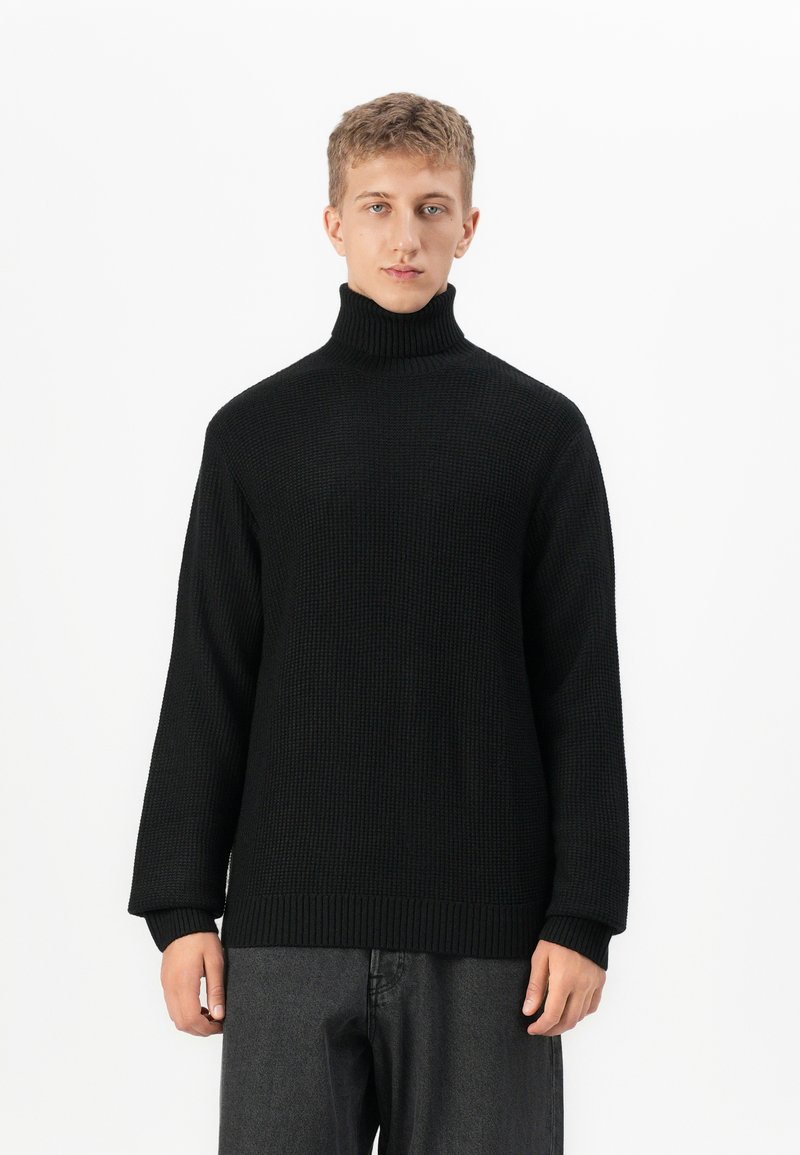 Jongen met kort haar, gekleed in een zwarte gebreide coltrui en donkergrijze broek, staand tegen een witte achtergrond.