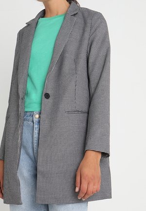 Vrouw draagt een zwart-wit pied-de-poule blazer over een teal T-shirt en lichtblauwe jeans, staand tegen een witte achtergrond.
