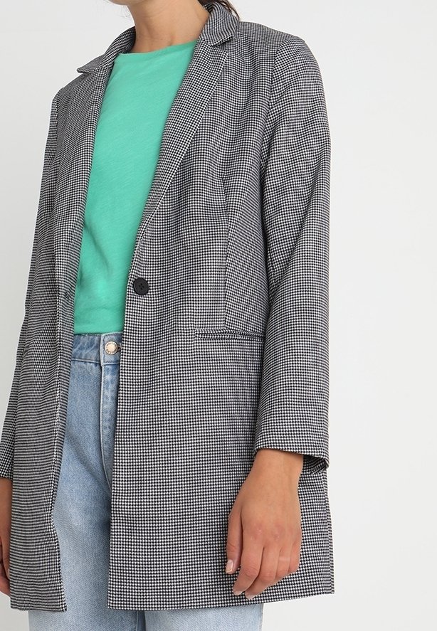 Femme portant un blazer à motif pied-de-poule noir et blanc sur une chemise bleu canard et un jean bleu clair, debout devant un fond blanc.