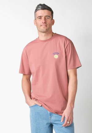 Muž nosiaci prachovo ružové tričko s malým žltým slnečným motívom a textom, v ľahko modrých džínsoch, stojaci pred jednoduchým svetlým pozadím.