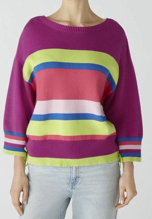 Person iført en flerfarvet stribet sweater med vide ærmer og lyseblå jeans, stående mod en ensfarvet baggrund.