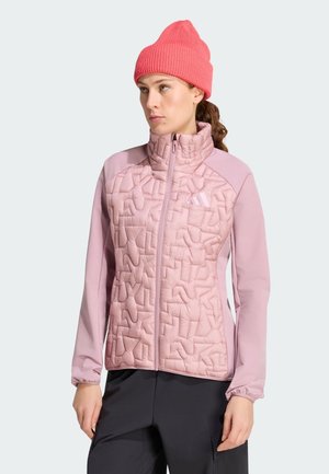 Vrouw draagt een roze doorgestikte jas met een hoge kraag, zwarte broek en een felroze gebreide muts, kijkt naar de zijkant tegen een effen achtergrond.