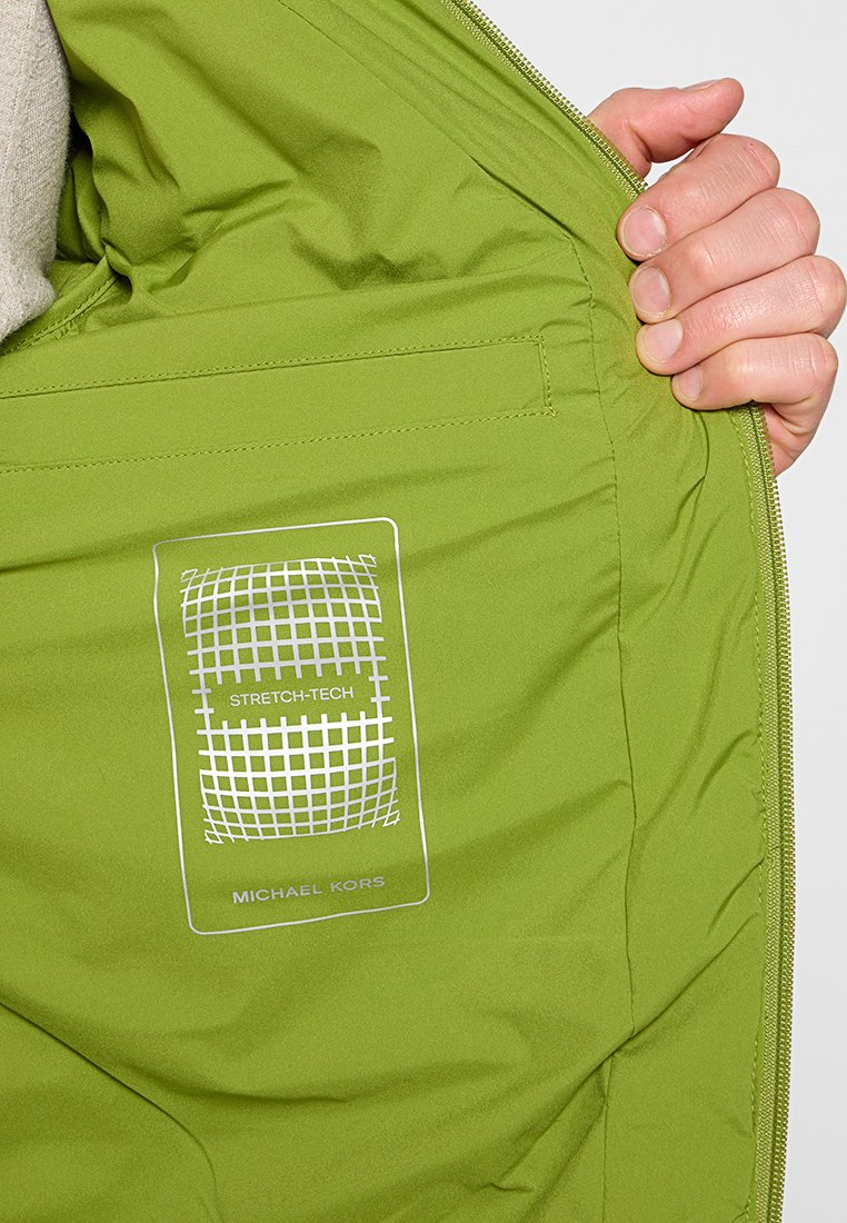 Interno della giacca verde che mostra una texture liscia e leggera. Presenta una tasca cucita e un'etichetta con "STRETCH-TECH" e "MICHAEL KORS."