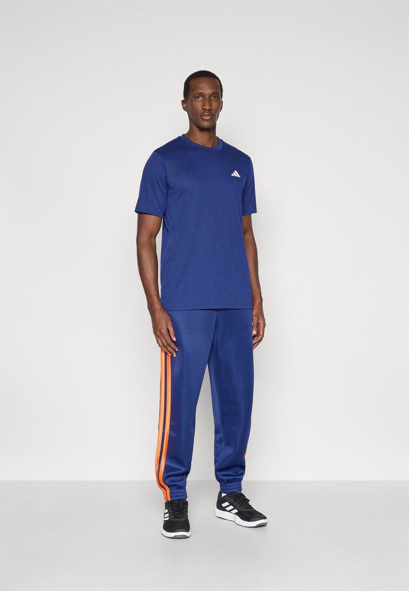 Marinefarbener sportlicher T-Shirt mit kurzen Ärmeln und einem kleinen weißen Logo auf der Brust, kombiniert mit marinefarbenen Jogginghosen mit orangen Streifen und elastischen Bündchen.