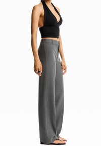 Femme portant un crop top noir sans manches à col halter, un pantalon gris taille haute à jambes larges, des bagues aux doigts et des sandales noires, debout de profil.