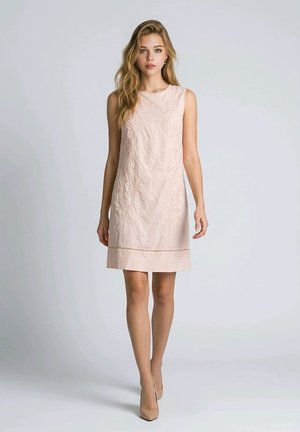 Joven mujer con un vestido sin mangas bordado de color rosa pálido y tacones beige caminando frente a un fondo liso de color gris claro.