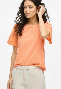 Camiseta color durazno hecha de tela suave, con detalle de encaje en las mangas, escote redondo y un corte relajado. Combinada con pantalones de color claro.