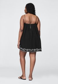 Črna obleka brez rokavov z zbiranimi detajli, okrašena z belim cvetličnim vezenjem na robu in petimi dekorativnimi gumbi na hrbtni strani.