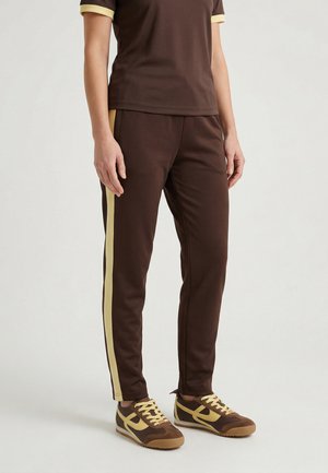 Osoba nosząca brązowe spodnie sportowe z beżowymi paskami po bokach oraz pasujące brązowo-beżowe sneakersy, stojąca na jednolitym tle.