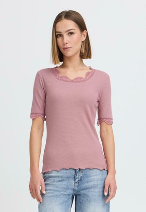 Giovane donna con capelli lisci castano chiaro che indossa una maglietta a maniche corte color malva con rifiniture in pizzo e jeans di denim azzurro chiaro.