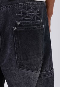Jeans in denim nero con un design unico cucito sulla tasca posteriore, dotati di due tasche inserite visibili e dettagli in metallo.