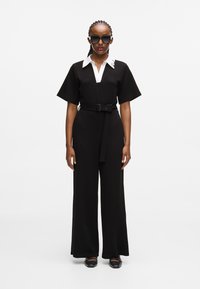 Čierny overal s bielym golierom, krátkymi rukávmi, opaskom v oblasti pása a širokými nohavicami. Modelka nosí prehnané čierne slnečné okuliare.
