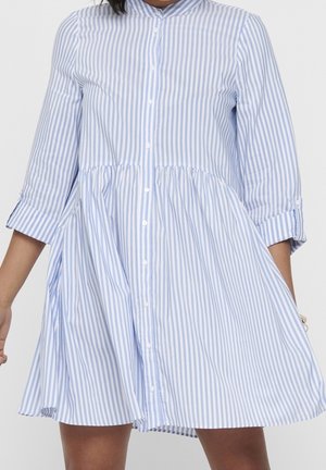 Vrouw draagt een blauw-wit verticale gestreepte blousejurk met driekwart mouwen en een losse, uitlopende rok.