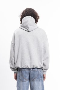 Persona con capelli ricci di spalle, che indossa un felpa grigia chiara e jeans blu sbiaditi su uno sfondo bianco.