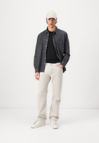 Dunkelgraues Hemd über einem schwarzen T-Shirt, hellbeige Jeans und weiße Sneaker. Das Model trägt eine beige Mütze. Einfaches, lässiges Outfit.