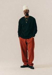 Uomo con un berretto crema, occhiali rotondi, maglione nero, collane d'oro, pantaloni color ruggine e scarpe nere, che sta in piedi con le mani in tasca.