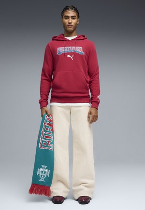 Joven con cabello trenzado que lleva una sudadera roja con la palabra "Portugal", pantalones anchos color crema, zapatos granate y sostiene una bufanda verde azulado de Portugal con flecos rojos.