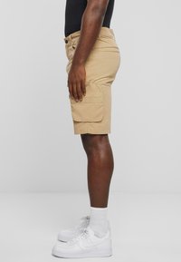 Shorts cargo beige en coton, dotés de poches latérales, d'une coupe droite et d'une fermeture à bouton, portés avec des baskets blanches et des chaussettes montant au mollet.