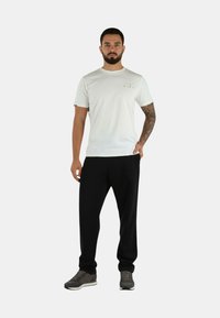 Maglietta bianca a maniche corte con un logo sul petto, abbinata a pantaloni neri a gamba stretta e sneakers grigie, in piedi su uno sfondo semplice.