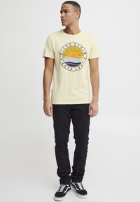 Gul bomulls t-skjorte med et rundt grafisk design av en solnedgang og bølger, kombinert med svarte denimbukser og svart-hvite sneakers.