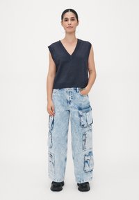Marinblå ärmlös V-ringad stickad topp som bärs med ljusblå, oversized cargojeans med flera fickor och en blekt tvätt, tillsammans med svarta skor.