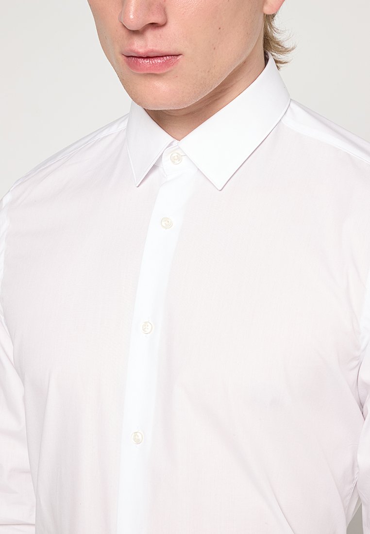 Camisa blanca de botones con un cuello clásico, mangas largas y una tela suave con una ligera textura. Siete botones en la parte delantera.