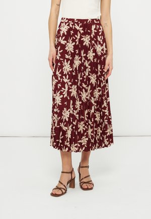 Ženska, oblečena v bordo plisirano midi krilo z bež cvetličnim potiskom, belo majico in rjave sandale s peto, stoji pred enobarvnim ozadjem.