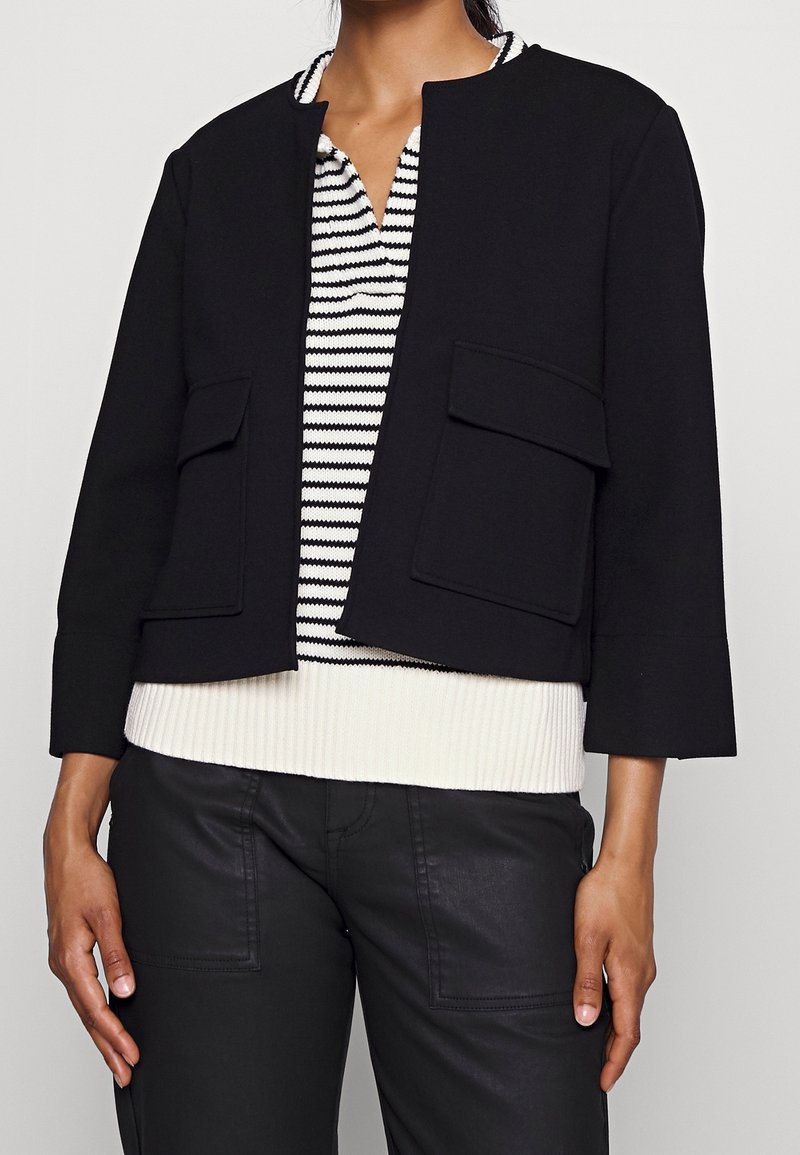 Femme portant une veste courte noire avec de grandes poches, un pull rayé noir et blanc en dessous, et un pantalon noir.