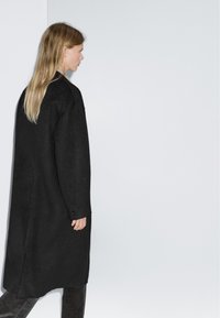 Cappotto in lana nero, oversize, con maniche lunghe, silhouette dritta, texture sottile e dettaglio con un solo bottone al polsino.