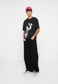 Czarny t-shirt graficzny z białym nadrukiem przedstawiającym osobę trzymającą mikrofon, w zestawieniu z luźnymi czarnymi spodniami i trampkami z niebieskimi akcentami.