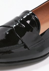 Mocassin en cuir verni noir avec talon bas, découpe décorative sur le dessus, et doublure intérieure beige, présenté en gros plan.