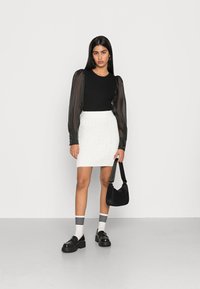 Top preto com textura em relevo e mangas bufantes transparentes, saia mini em tricot branco, mala preta, sapatos pretos chunky e meias de tornozelo listradas.